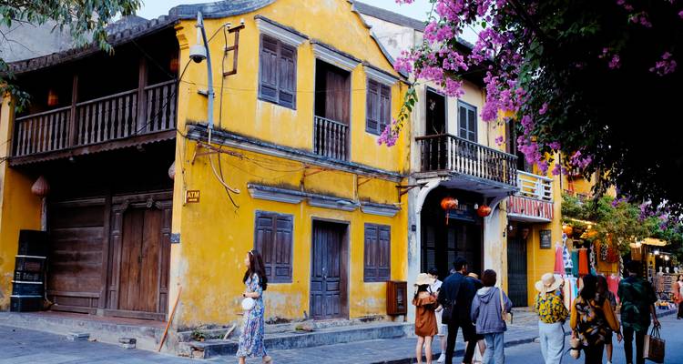 Des bâtiments patrimoniaux colorés et des bougainvilliers bordent une ruelle de Hoi An tandis que des touristes se promènent en dessous.