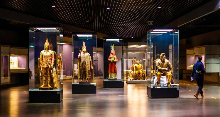 Sala de museo moderna que exhibe armadura de guerrero dorada y figuras históricas en vitrinas bajo una iluminación dramática.