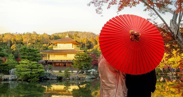 Twee bezoekers in kimono staan bij een vijver met een opvallende rode parasol en kijken naar Kyoto's gouden Kinkaku-ji die weerkaatst in het water.