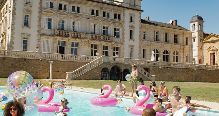 Fête de piscine animée dans un grand château français avec des bouées flamants roses et des baigneurs de soleil.