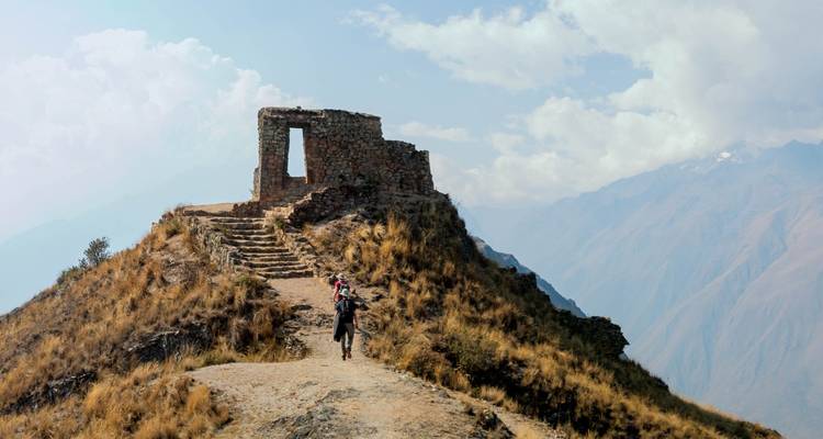 Wandelaar die een kleine stenen ruïne nadert bovenop een dramatische bergkam onder heldere luchten.