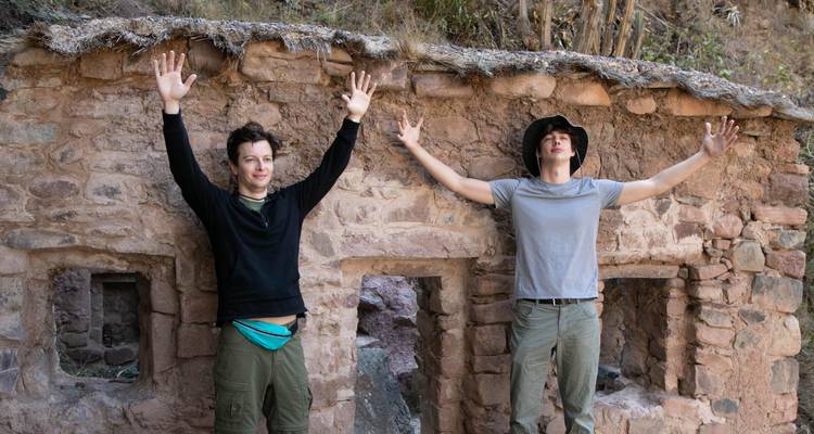 Twee trekkers steken hun armen omhoog tegen een rustieke stenen muur van oude ruïnes.