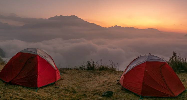 Twee rode tenten opgezet boven een zee van wolken bij dageraad met bergsilhouetten in de verte.