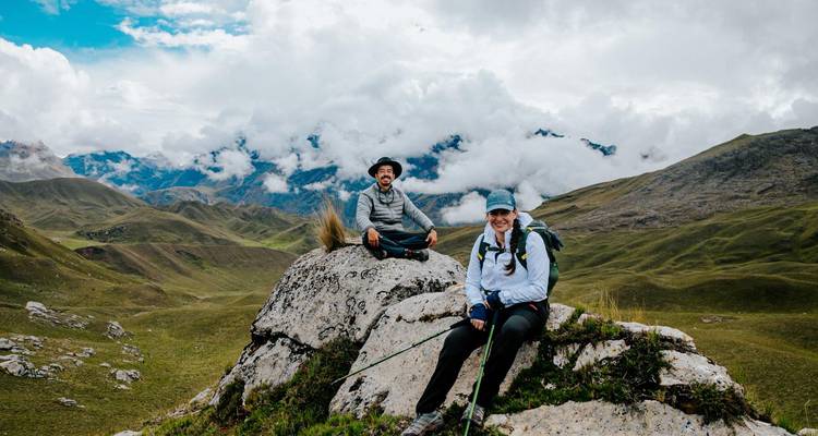 Twee trekkers rusten op een rotsachtige uitstulping te midden van een uitgestrekt berglandschap bedekt met wolken.