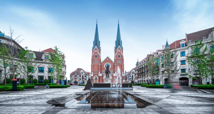 Place moderne symétrique avec église en brique rouge à deux clochers et fontaine réfléchissante au premier plan.