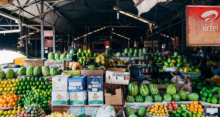Étals colorés chargés de fruits à l'intérieur d'un marché couvert avec des caisses, des melons et des agrumes empilés en hauteur.