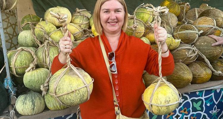 Glimlachende vrouw op een markt houdt twee bundels meloenen vast voor een kraam vol met pompoenen.
