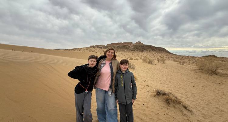 Een vrouw en twee kinderen staan in zandig woestijnterrein met een vestingruïne in de verte onder bewolkte luchten.