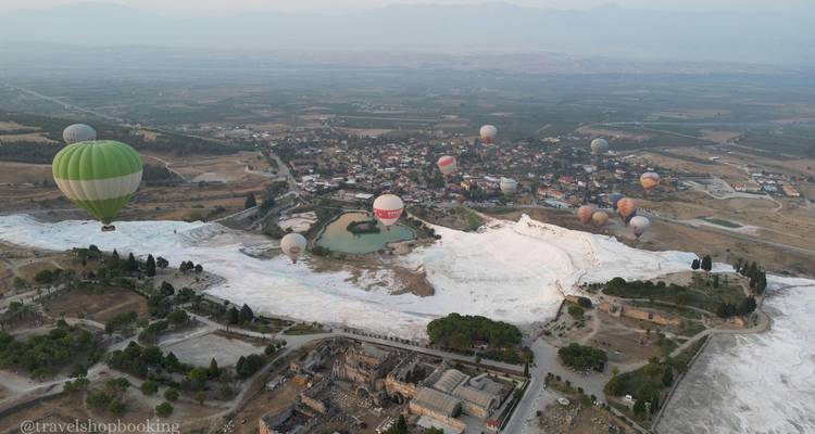 Vista aérea de las terrazas de travertino blanco de Pamukkale con numerosos globos aerostáticos coloridos flotando sobre un pequeño pueblo y tierras de cultivo circundantes.