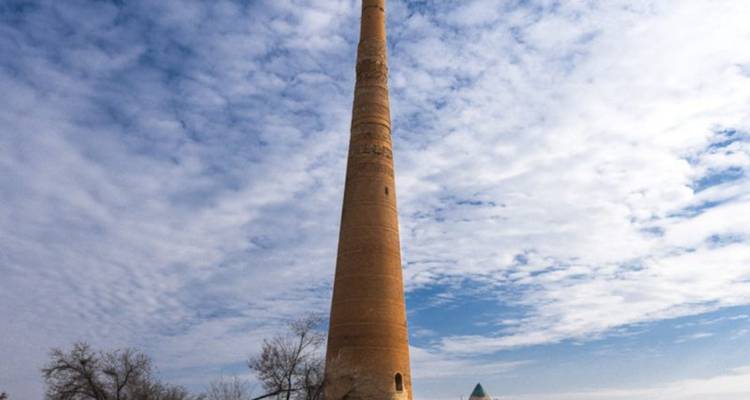 Hoge slanke bakstenen minaret die oprijst in een gedeeltelijk bewolkte hemel met kaal land eromheen.