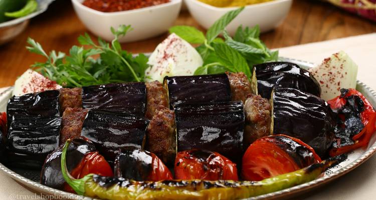 Primer plano de un plato de kebab turco con berenjena asada, tomates y hierbas.