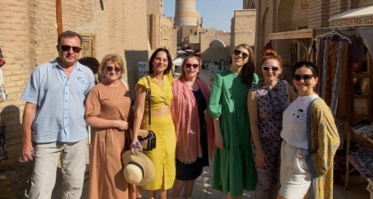 Zeven reizigers poseren in een door de zon verlichte steeg van een oude woestijnstad met de turkoois betegelde Kalta Minor minaret en modderblokken muren achter hen.