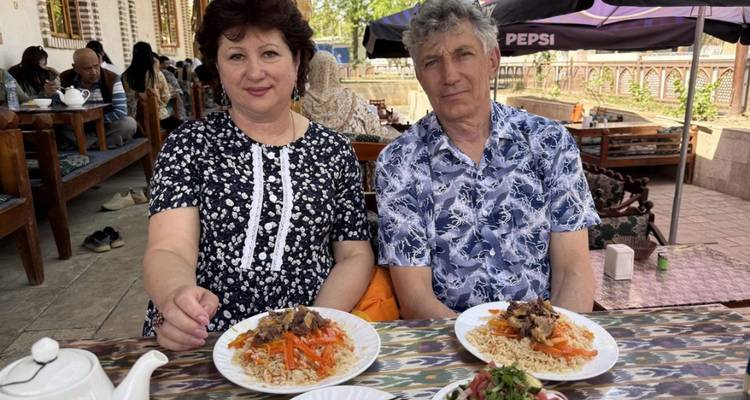 Een ouder echtpaar zit aan een tafel van een buitenrestaurant en geniet van borden met Oezbeekse plov en salade onder een Pepsi-parasol.