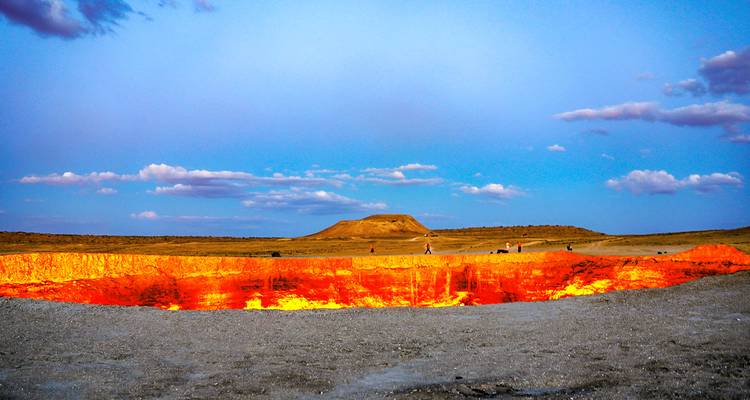 Vista amplia diurna del cráter de Darvaza brillando en color naranja bajo un cielo azul brillante y llanuras abiertas.