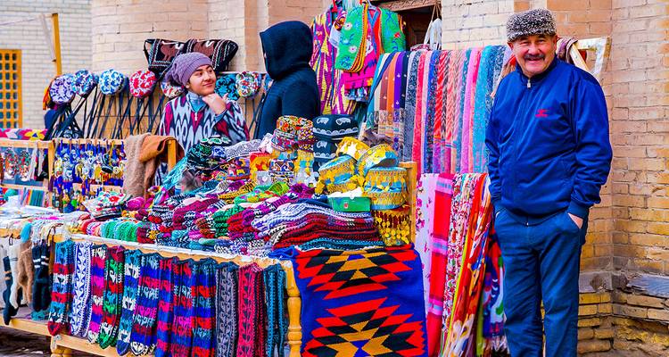 Puesto de bazar colorido apilado con bufandas tejidas, sombreros y alfombras, atendido por un vendedor sonriente.