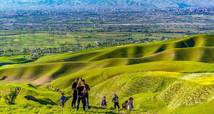 Las familias caminan por colinas verdes ondulantes con vista a una ciudad extensa y montañas distantes.