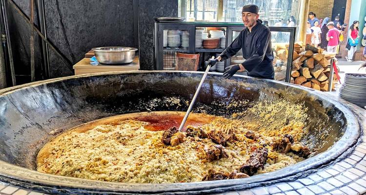 Le chef remue un immense chaudron de riz et de viande plov fumants à un étal alimentaire extérieur tandis que les visiteurs regardent.