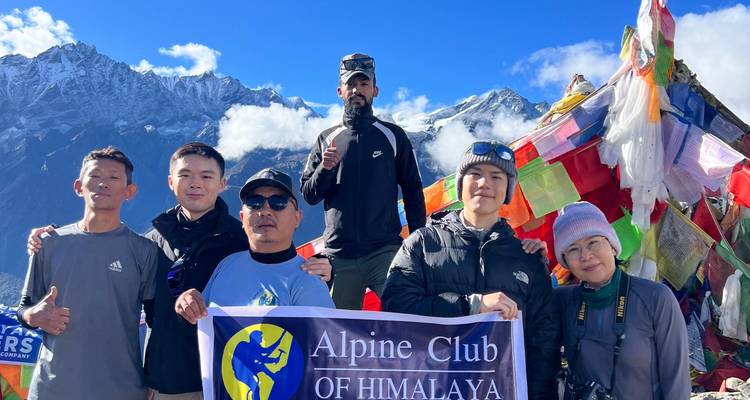 Wanderer posieren an einem mit Gebetsfahnen bedeckten Aussichtspunkt mit schroffen Himalaya-Gipfeln und blauem Himmel.