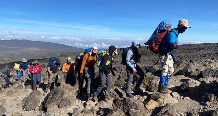 Un groupe de randonneurs gravit un sentier rocheux en altitude sur le mont Kilimandjaro sous un ciel bleu dégagé.