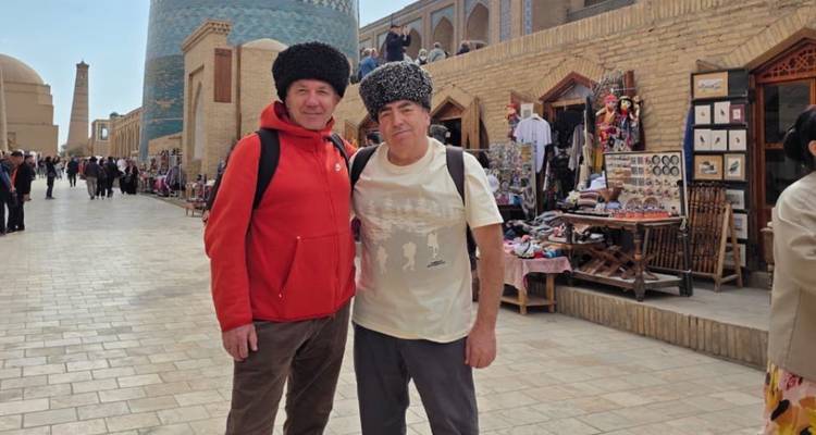 Twee mannen met traditionele bontmutsen poseren op een betegelde straat met Khiva's blauwe Kalta-Minor minaret achter hen.
