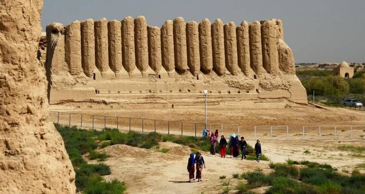 Bezoekers trekken langs een zanderig pad naar massieve geribbelde modderen muren van een oud fort te midden van schaarse woestijnvegetatie.