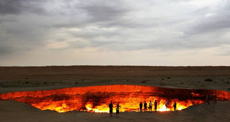 Gesilhouetteerde reizigers staan aan de rand van de gloeiende Darvaza gaskrater onder een dramatische bewolkte hemel.