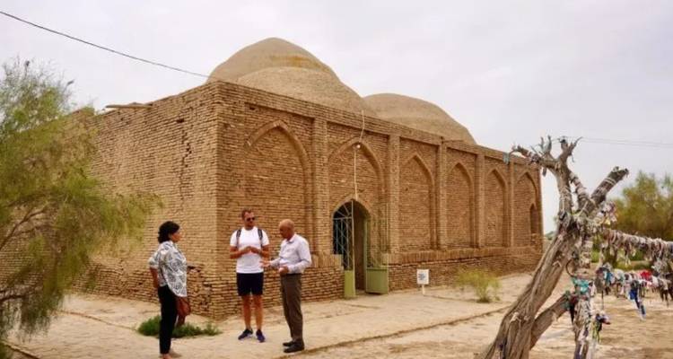 Drie bezoekers praten buiten een klein bakstenen koepelmausoleum in een stoffige binnenplaats.