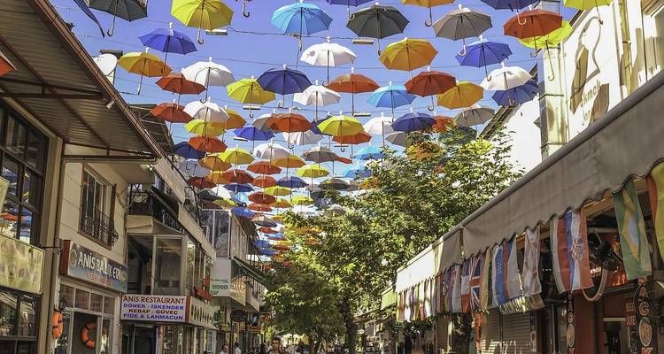 Levendige voetgangersstraat omzoomd met winkels en restaurants onder een kleurrijk baldakijn van hangende paraplu's.