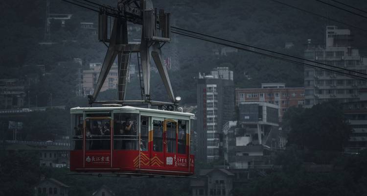 Rode stedelijke kabelbaan cabine glijdend aan kabels boven een somber stadsbeeld van grijze hoogbouw en heuvels.