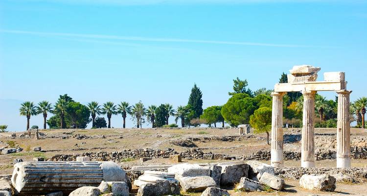 Zongebleekte archeologische ruïnes met staande zuilen en verspreide stenen blokken onder een heldere blauwe hemel.