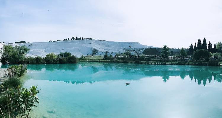 Turquoise thermale poel die witte kalkstenen terrassen en groen in de verte weerkaatst op een heldere dag.