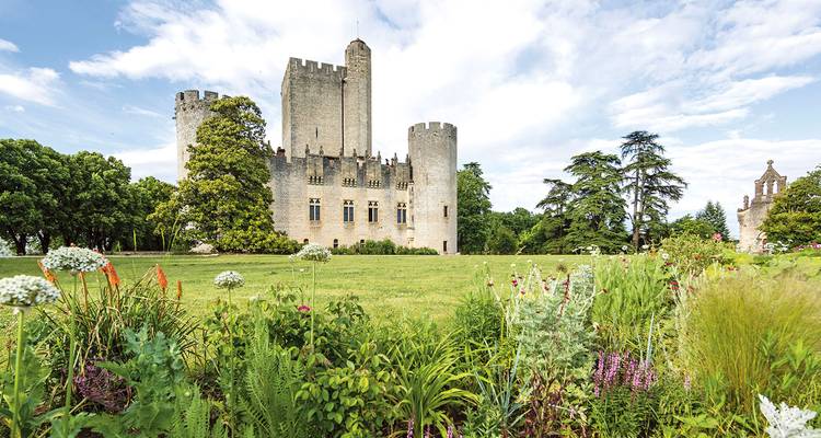 Château de pierre médiéval avec tours rondes situé derrière un jardin luxuriant et une large pelouse sous un ciel partiellement nuageux.