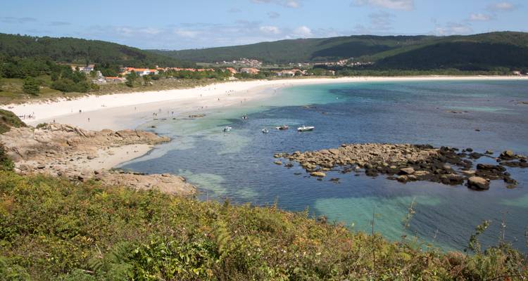 Agua turquesa cristalina y playa arenosa con pequeñas embarcaciones y pueblo distante respaldado por colinas verdes