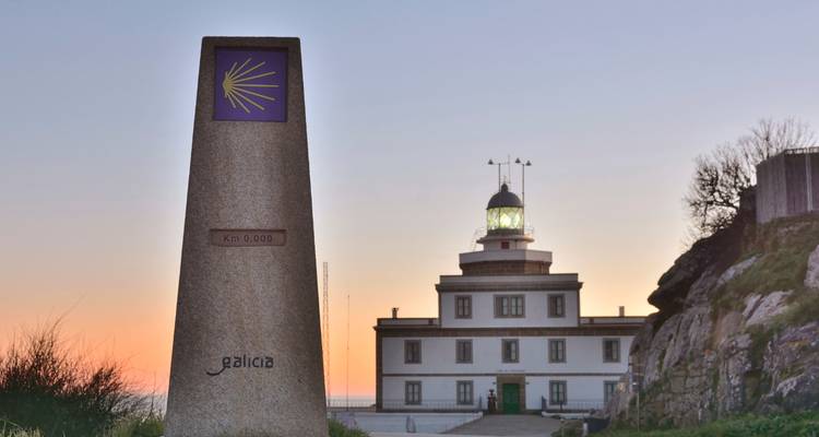 Faro de Finisterre y marcador del kilómetro cero del Camino recortados contra un cielo de atardecer colorido