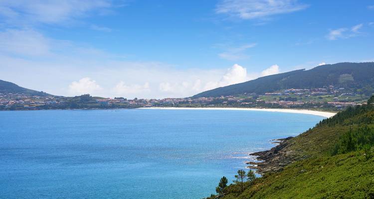 Amplia bahía azul bordeada por colinas verdes y pueblo costero distante bajo cielos brillantes