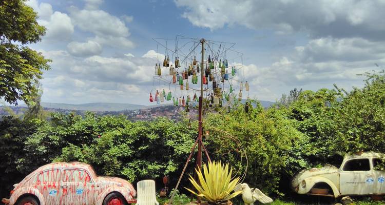 Installation artistique extérieure composée de bouteilles suspendues sur une structure métallique entre deux voitures vintage éclaboussées de peinture et une végétation luxuriante.