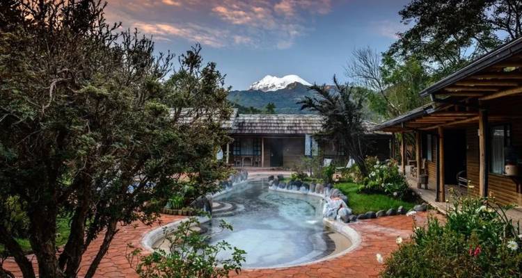 Malerischer Spa-Pool mit aufsteigendem Dampf, üppigen Gärten und schneebedeckter Bergkulisse bei Sonnenuntergang.