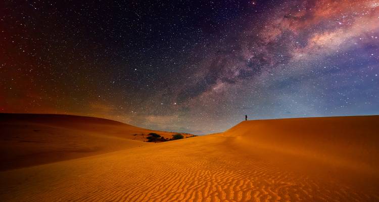 Cielo nocturno lleno de estrellas y arco de la Vía Láctea sobre dunas doradas del desierto con una figura solitaria.