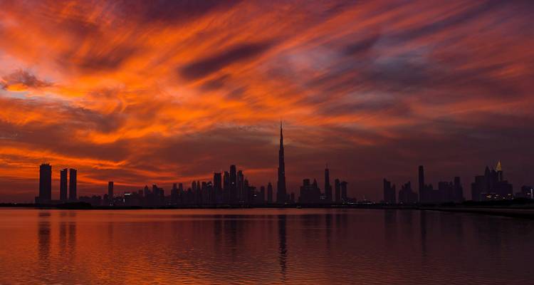 Cielos de atardecer naranja ardiente sobre el perfil silueteado de Dubai reflejado en aguas tranquilas.