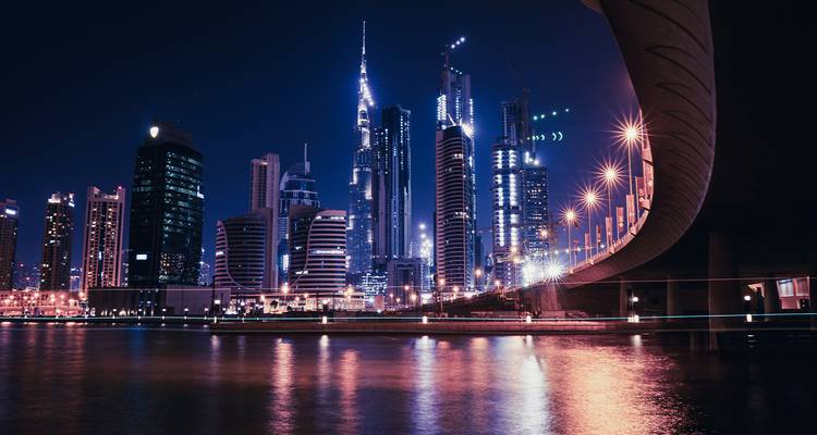 Vista nocturna desde la orilla del río de los rascacielos iluminados de Dubái con reflejos en el agua y un puente curvado.
