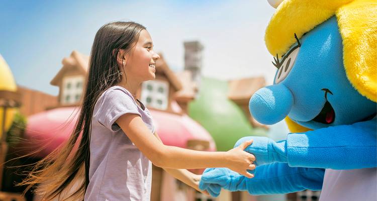 Niña sonriente juguetonamente tomada de la mano con una mascota de personaje de dibujos animados azul en un parque temático.