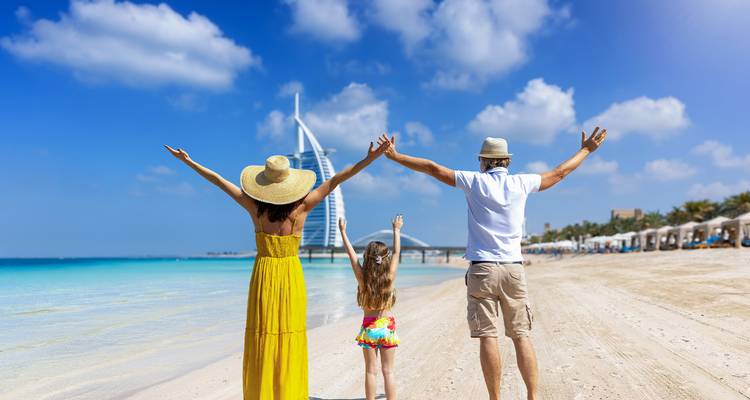 Familia con brazos alzados frente al mar turquesa y el Burj Al Arab en una playa soleada.