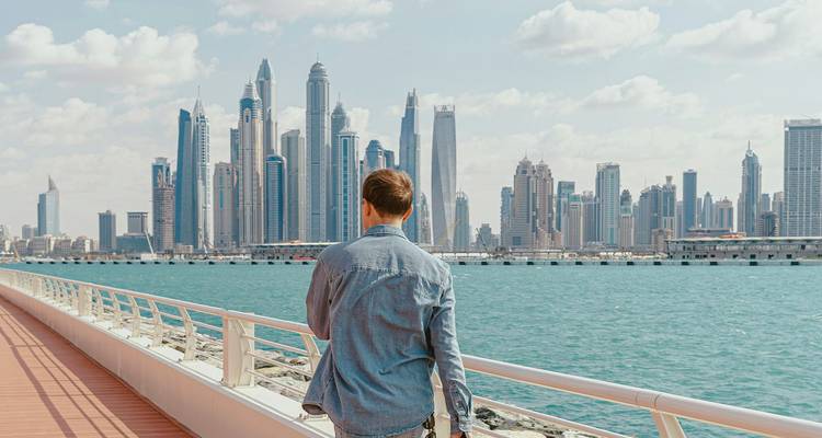 Un homme vêtu d'une veste en denim marche le long d'une promenade moderne surplombant l'eau turquoise et la ligne d'horizon des gratte-ciel de la Marina de Dubaï.