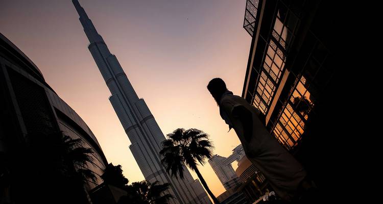 Silhouette d'une femme en tenue traditionnelle regardant vers le haut la Burj Khalifa alors que le ciel s'illumine d'une teinte orange au coucher du soleil.