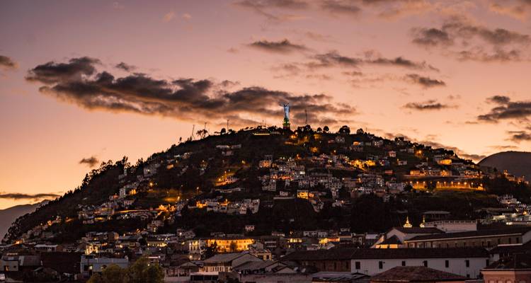Quito-Skyline bei Sonnenuntergang mit dem Panecillo-Hügel und der beleuchteten Jungfrauenstatue unter leuchtenden Wolken.
