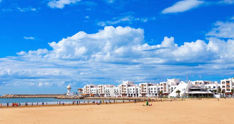 Breed zandstrand in Agadir omzoomd met witte kustgebouwen en verspreide bezoekers onder pluizige wolken.