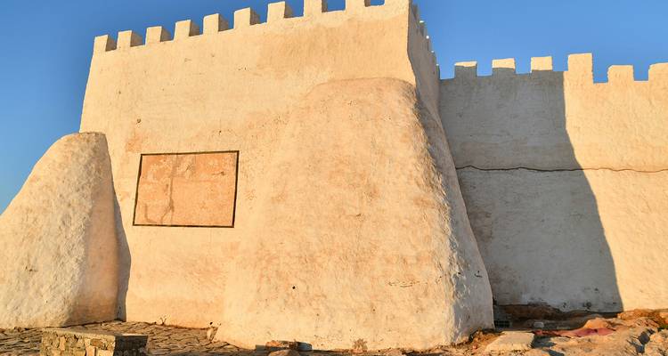 Eenvoudige door zonlicht beschenen adobe fortmuur met kantelen tegen een heldere blauwe lucht.