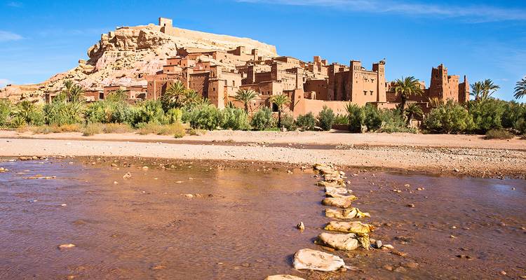 Fortaleza histórica de adobe de Ait Benhaddou que se alza sobre un lecho de río poco profundo con cruce de piedras bajo un cielo azul despejado.