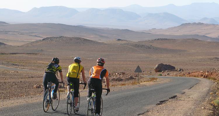 Des cyclistes pédalent le long d'une route sinueuse avec de vastes montagnes arides qui s'estompent dans la brume au loin.