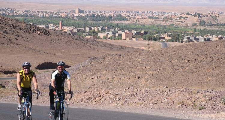 Deux cyclistes gravissent une pente surplombant une vallée verdoyante parsemée de bâtiments de ville marocaine.